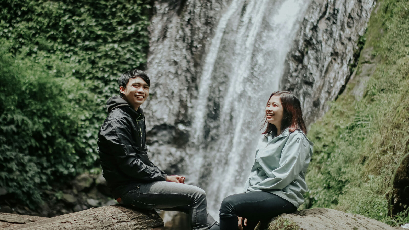 Man and woman are sitting on a rock in front of a waterfall.