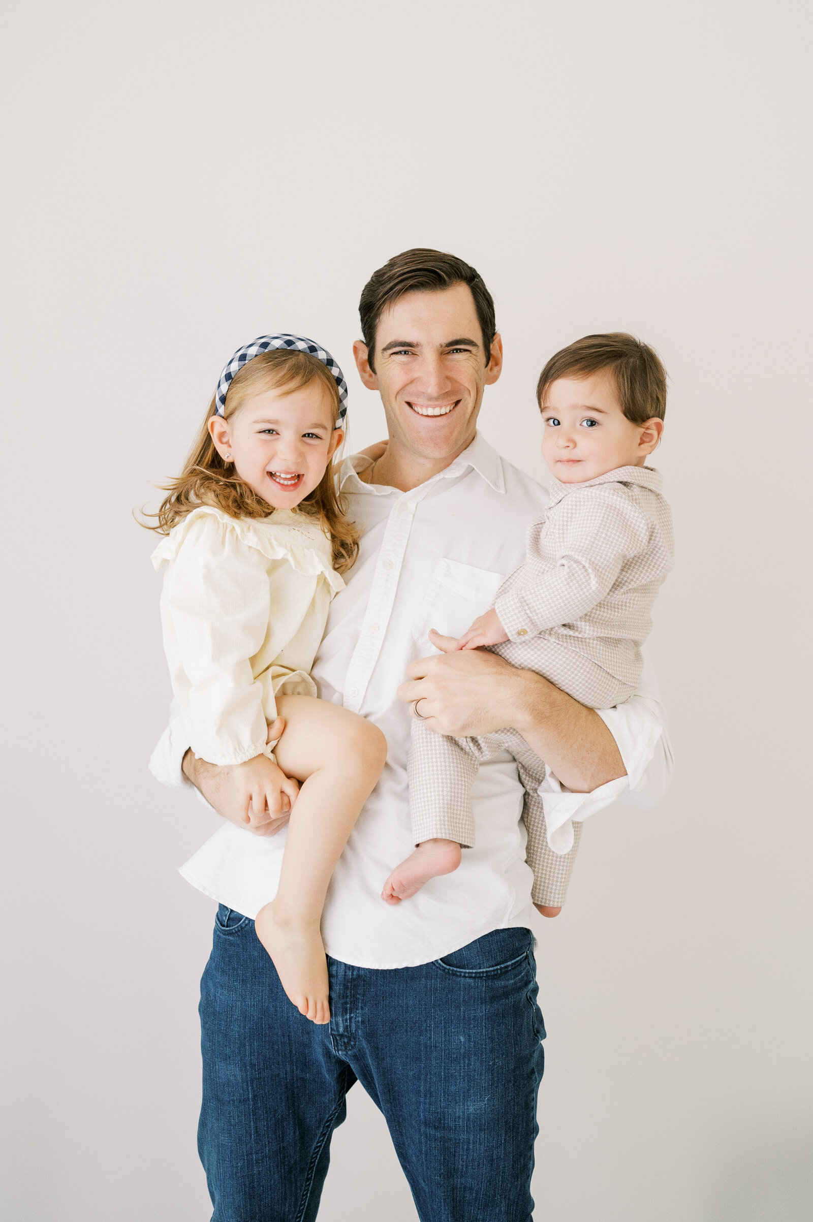 Dad holds young girl and toddler boy while smiling at the camera during studio family session in Raleigh, North Carolina