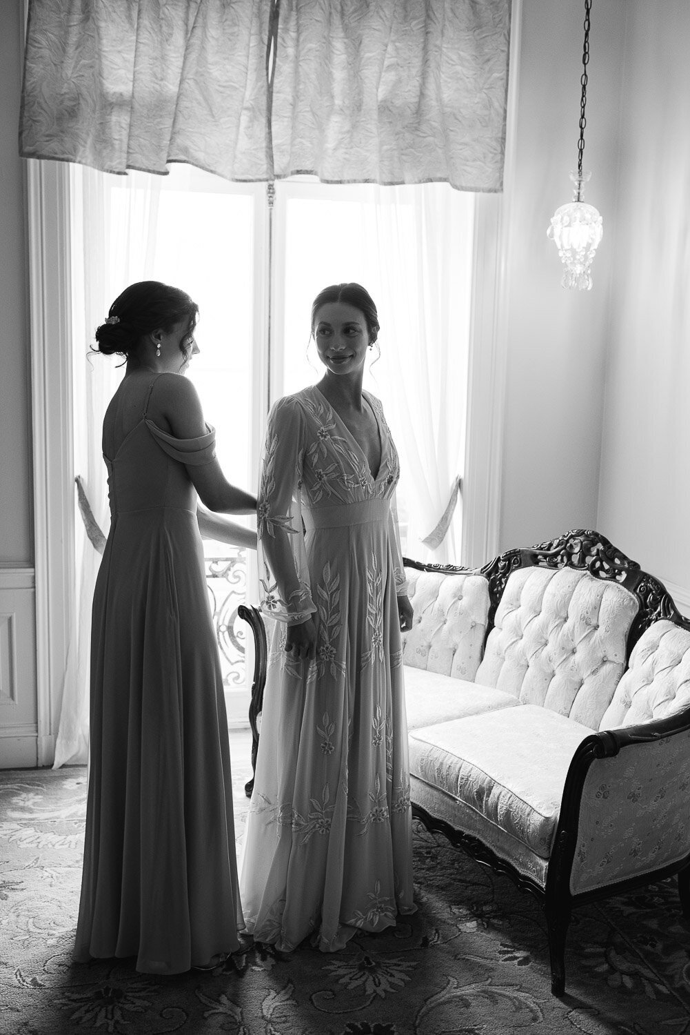 bridesmaid helps bride with dress in front of window