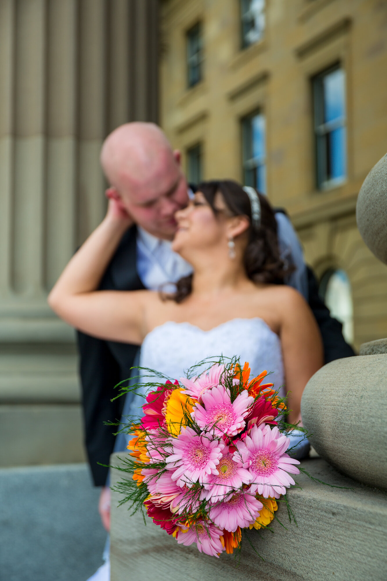 Bride leaning back arm around grooms head pulling him inat steps of Alberta Legislative Grounds