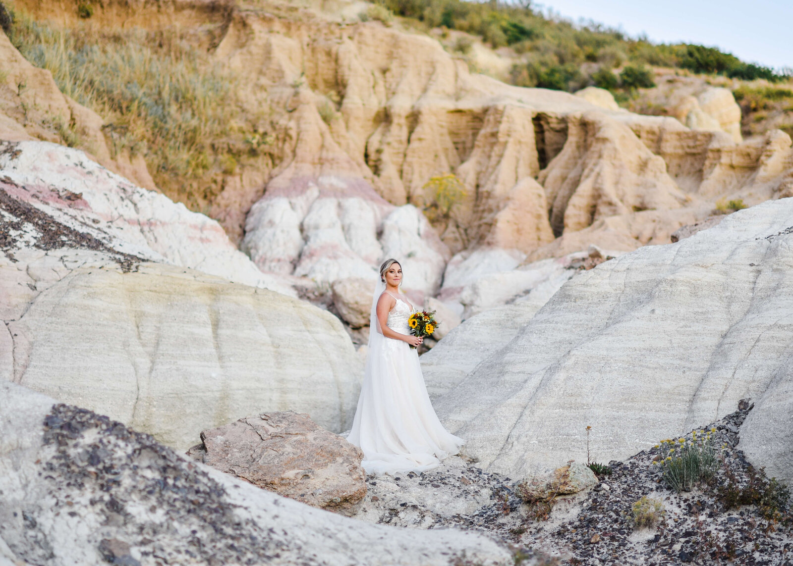 Bride in a white flowing gown holds flowers and stands on a picturesque rock