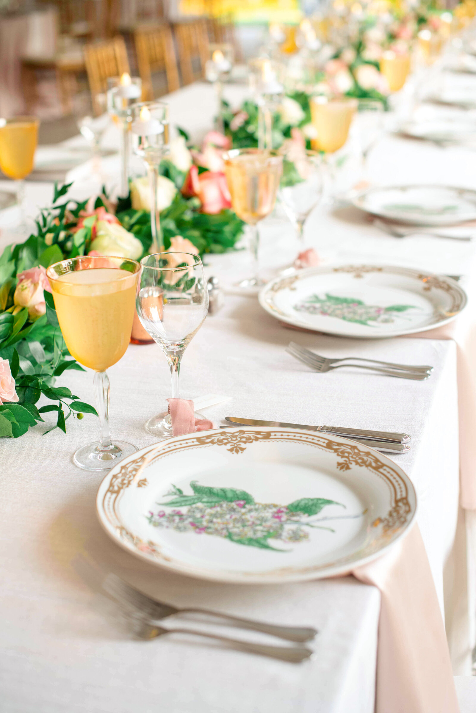 A long table is elegantly set with white plates featuring floral designs, gold-rimmed glasses, and silver cutlery. Greenery and pink flowers are arranged as a centerpiece, creating a sophisticated and inviting atmosphere.
