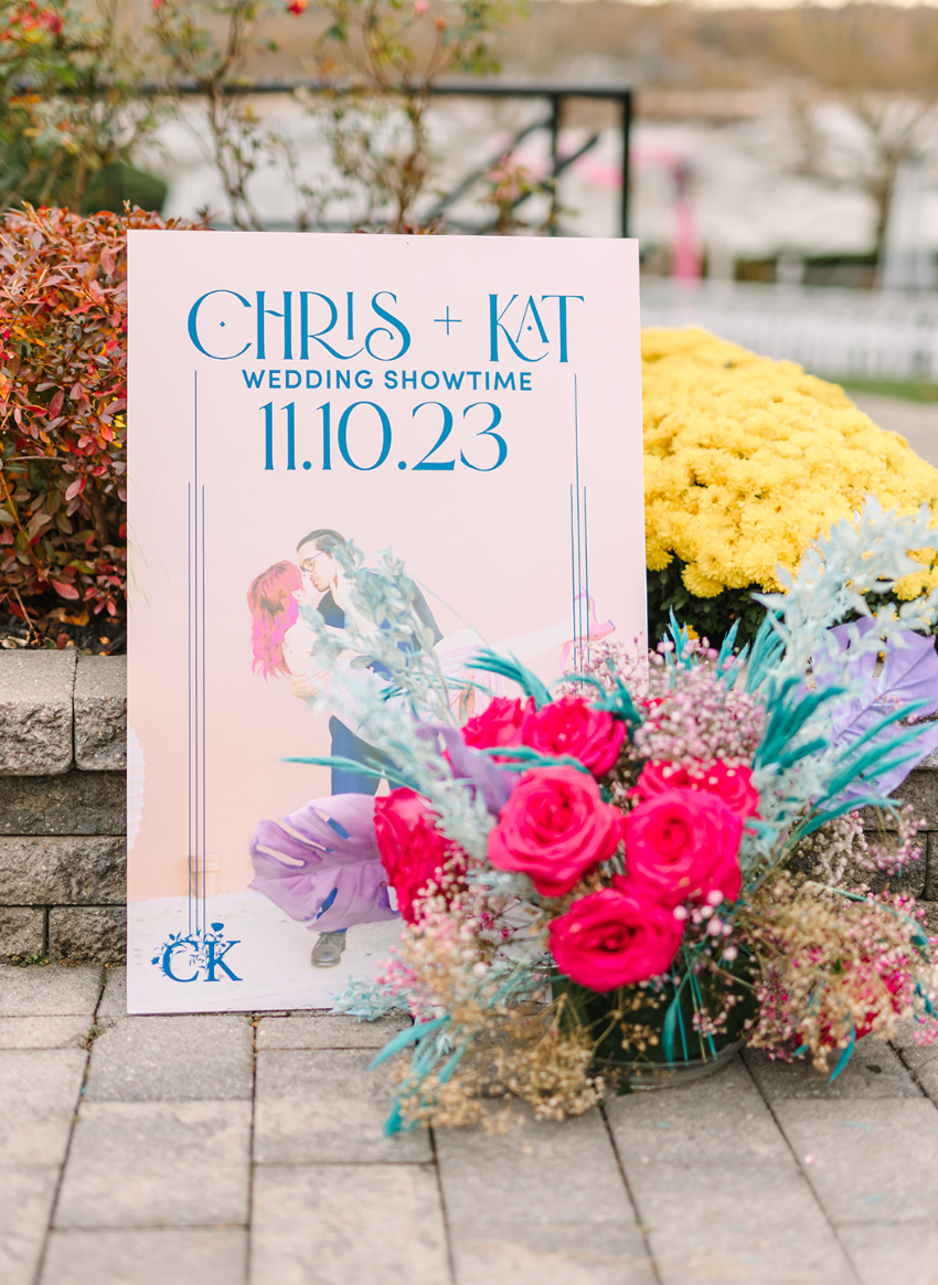 A wedding welcome sign showing a engaged couple kissing with colorful flowers in front of the welcome sign.