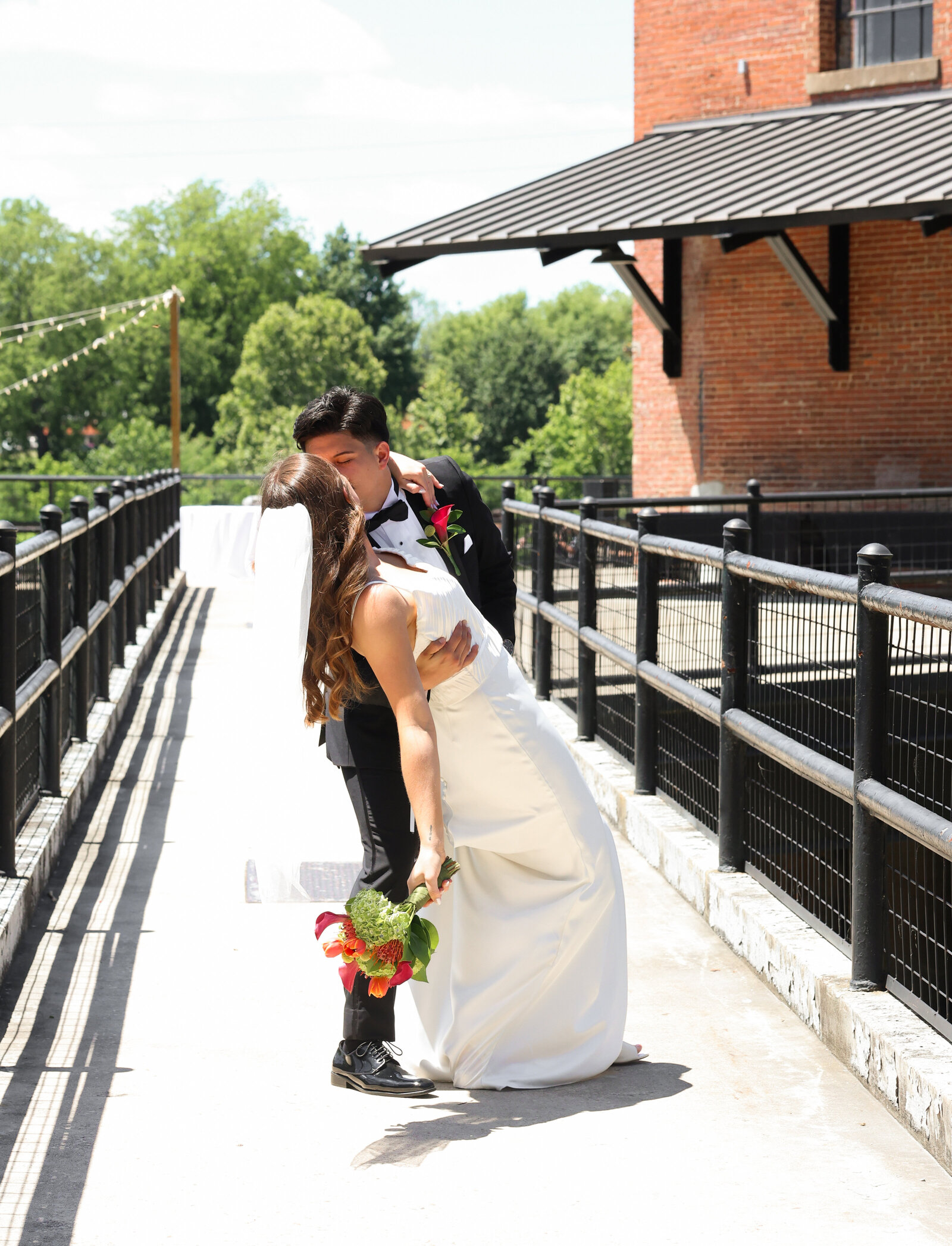 bride wearing white silk wedding dress and groom wearing black tuxedo kissing on walkway