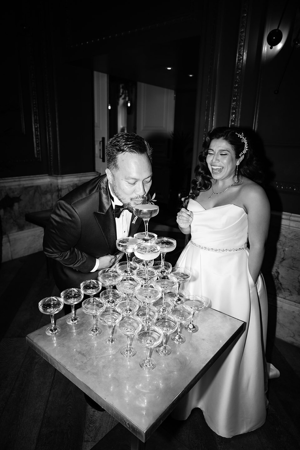 groom sipping on champagne next to laughing bride and champagne tower captured in editorial hard flash way