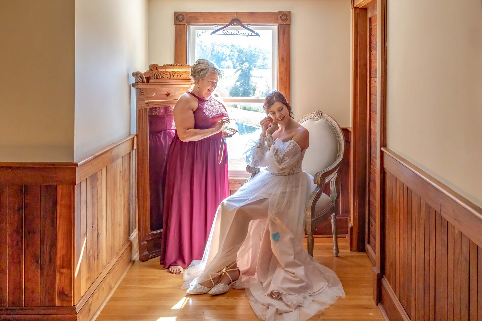 Bride sitting elegantly in a sunlit room as her mother helps her prepare for the wedding day, showcasing warmth and love in Apex, NC.
