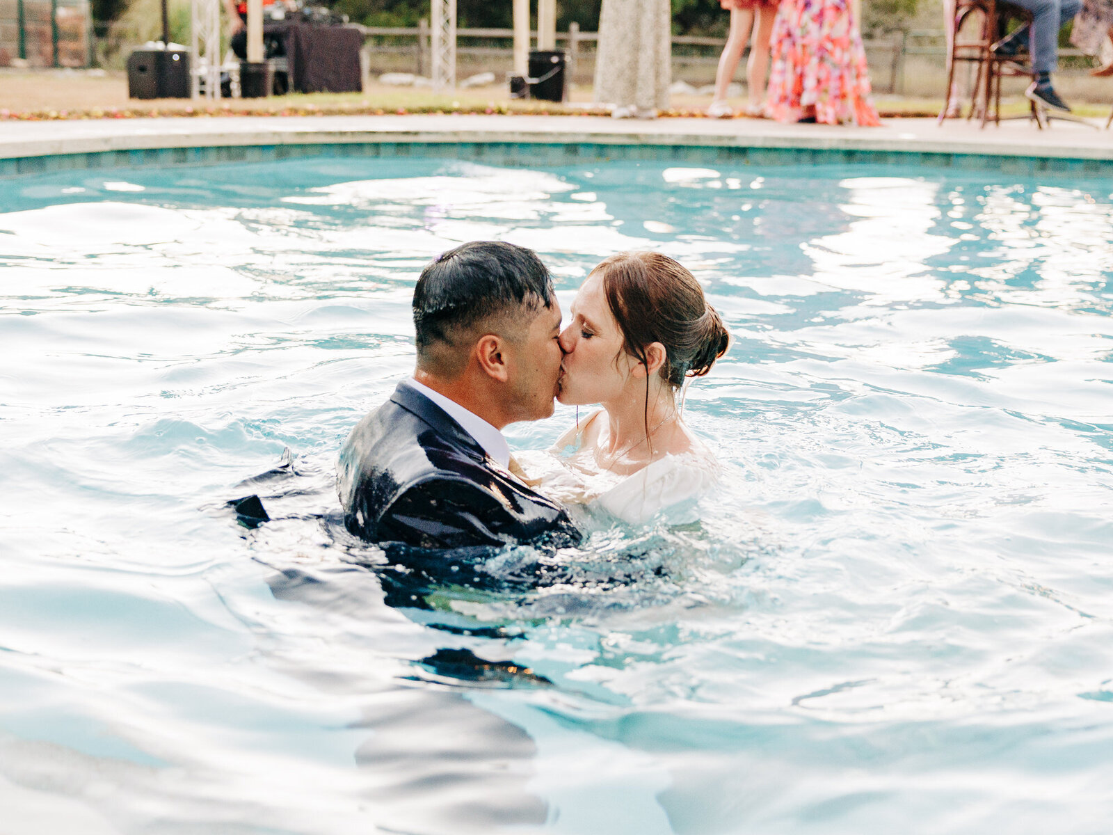 A bride and groom, dressed in wedding attire, passionately kiss while standing together in a swimming pool. Captured by a San Antonio Wedding Photographer, guests and beautiful wedding decorations are visible in the background.