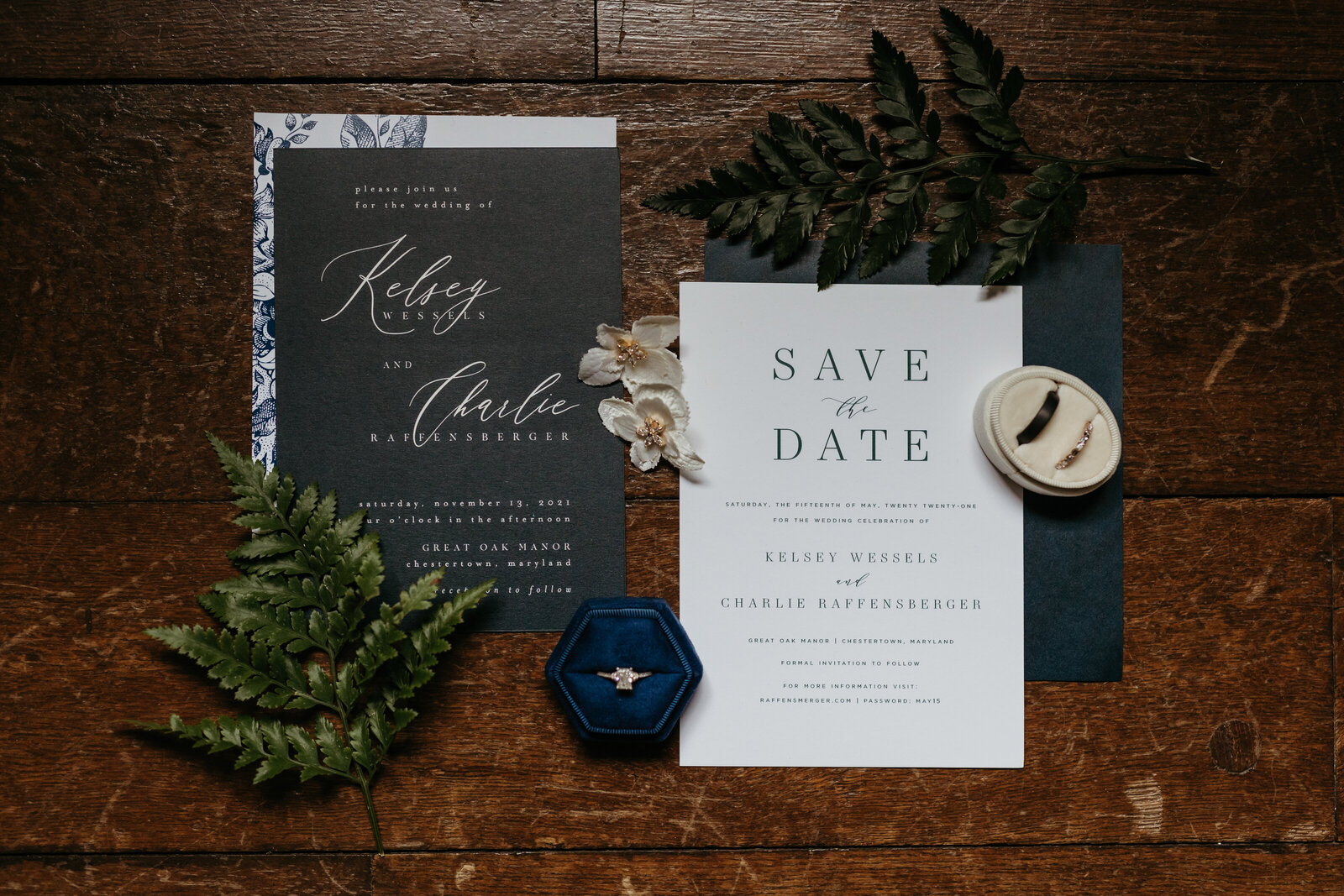 A suite of wedding invitations sits on a wood floor with ferns and wedding rings