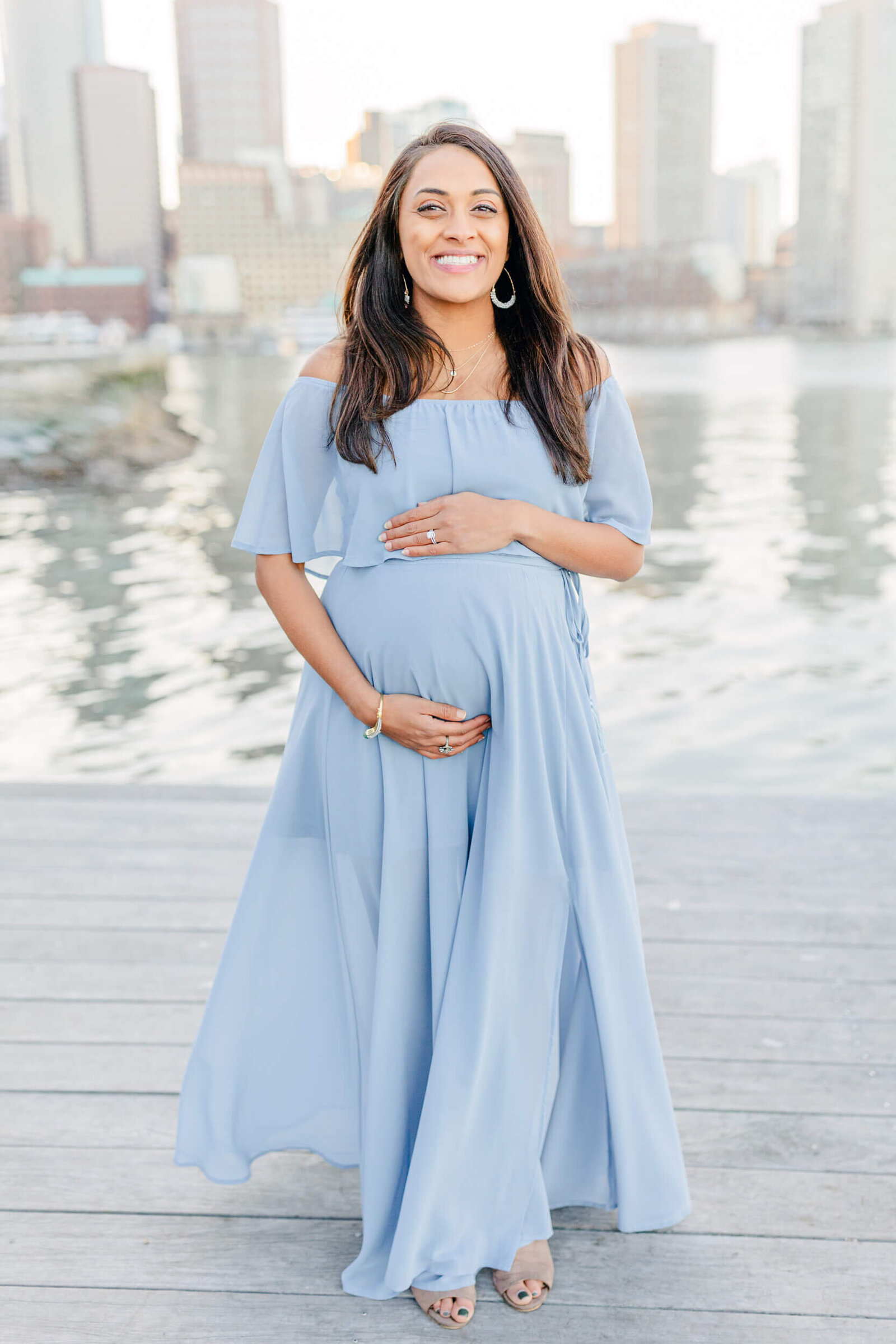 A pregnant woman smiles glowingly while holding her baby bump during a session with a Boston Maternity Photographer