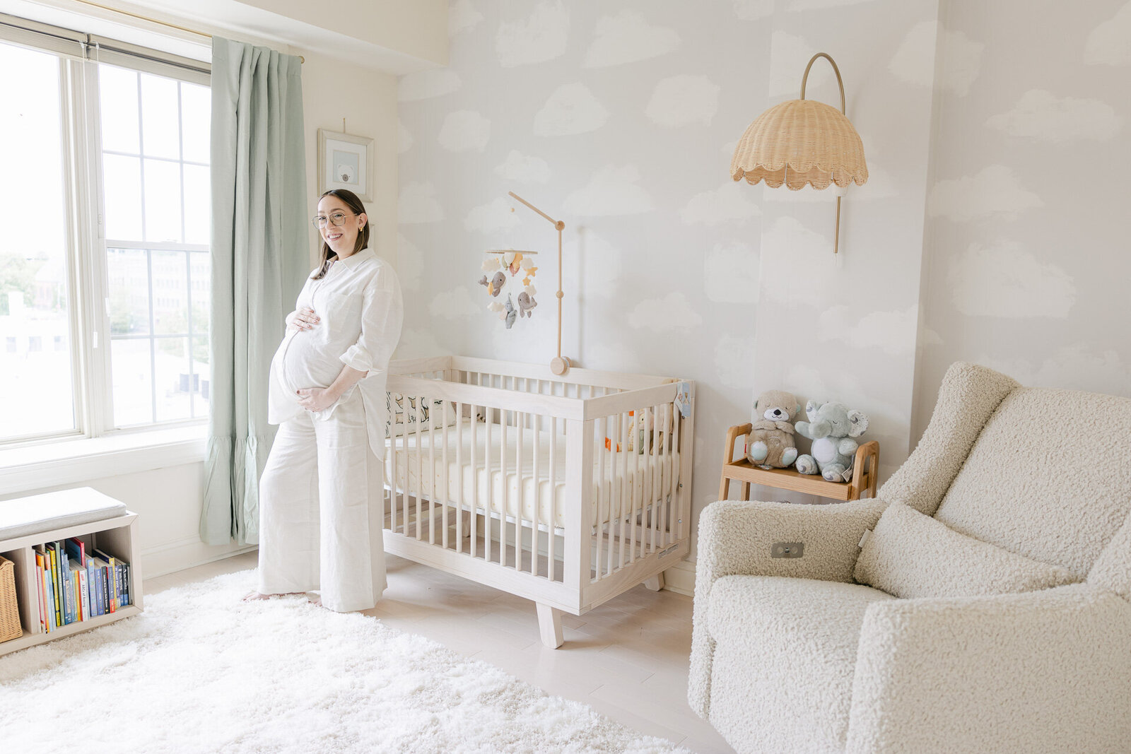 A glowing mom-to-be stands peacefully in a dreamy, cloud-themed nursery filled with soft textures and neutral tones. She cradles her bump next to a cream-colored crib, surrounded by light streaming through the window and plush toys nearby. The photo captures the cozy, calm energy of preparing for baby and makes the perfect inspiration for your own maternity session setup.