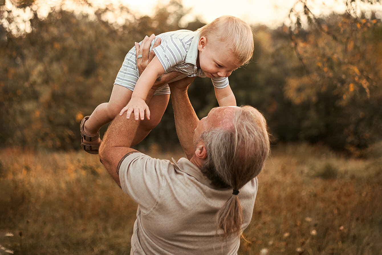 In the middle of a meadow a father and son. Dad is standing with the back to the camera and lifting the little boy above his head. The little boy is smiling at dad. Dad and me portrait. Family and Children Photography.