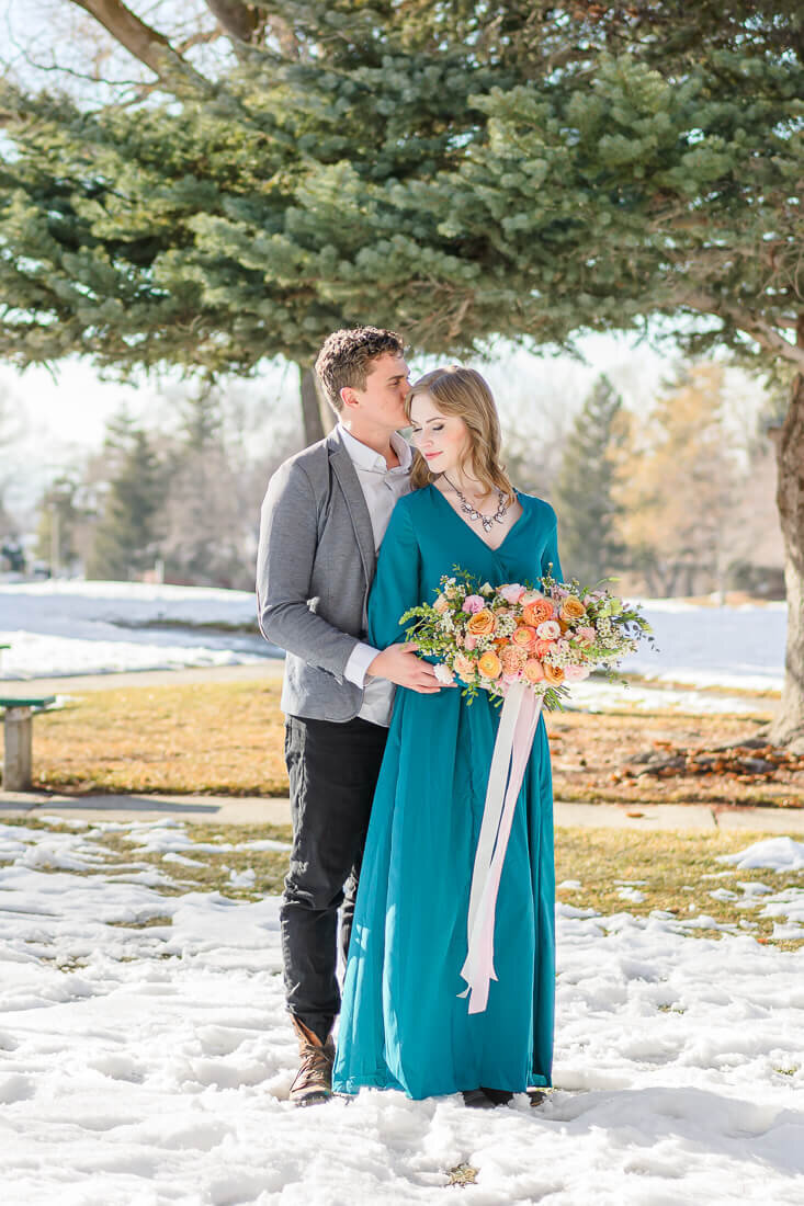 Utah engagement photography of a man in a white button-up shirt and a gray blazer standing behind his his fiancee wearing a long sleeved dark teal dress, holding a bouquet of orange and pink roses while standing in a park in Salt Lake City