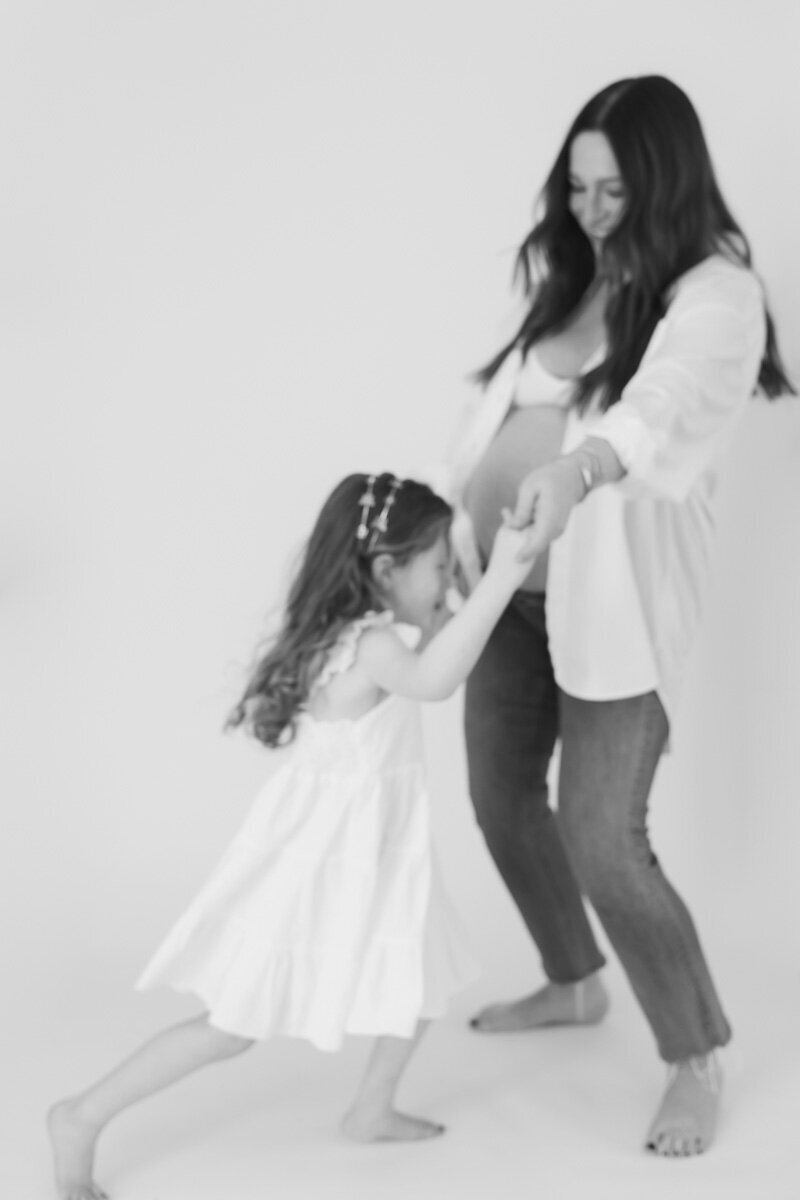 A tender black-and-white maternity portrait featuring a pregnant mother holding hands with her young daughter, who gently kisses her baby bump. The mother wears a white button-up shirt over a bra with dark jeans, while the daughter wears a flowy white dress with pearl accents in her hair. A sweet, emotional moment highlighting connection and anticipation.