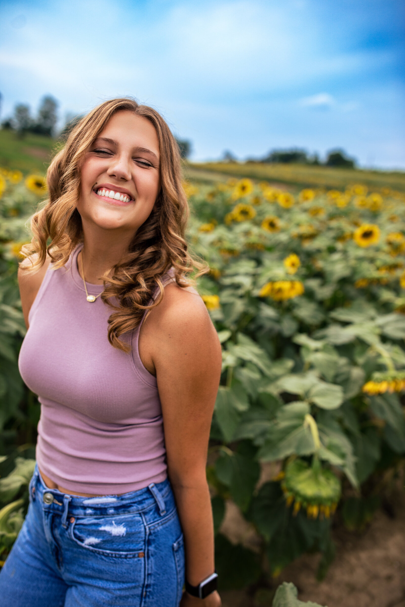 Say cheese! This cute senior smiles and squints up at the camera for her senior photo shoot in a field full of sunflowers.
