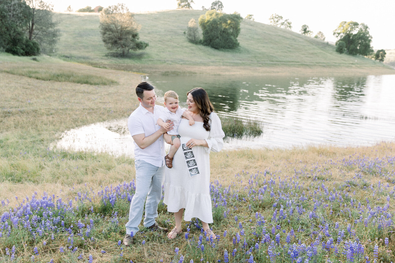 Image of expecting mother  holding an ultrasound image and big brother standing in wildflowers with father, taken by Sacramento Maternity Photographer Kelsey krall