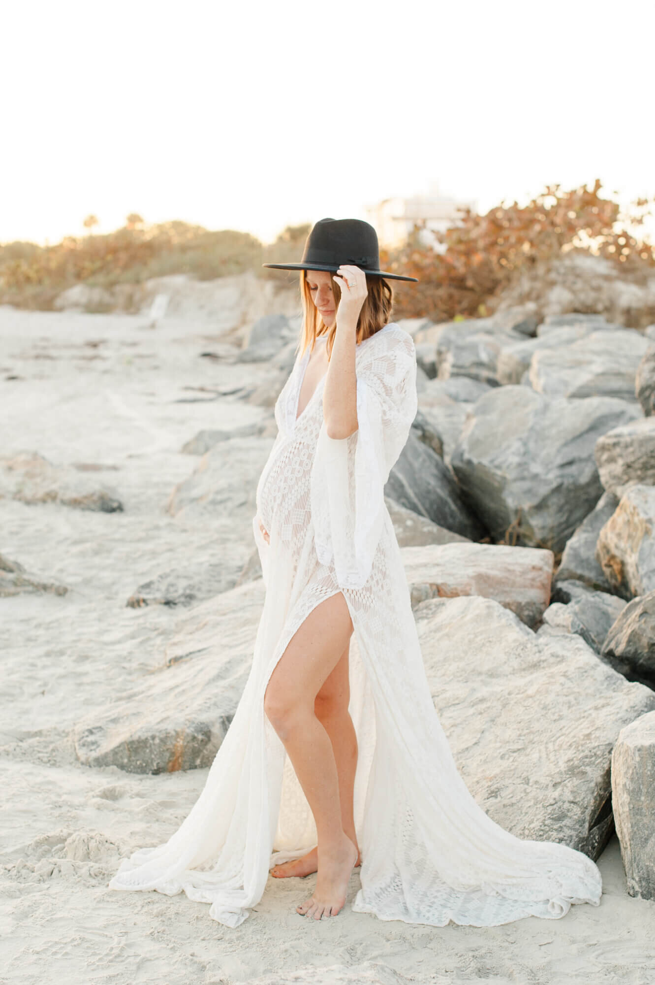 Pregnant mother stands near rocks on the beach holding her belly wearing a white lace gown