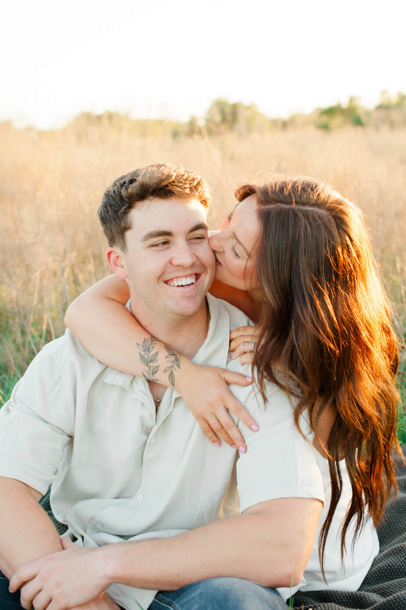 Couple smiles while kissing in a tall grass field at sunset