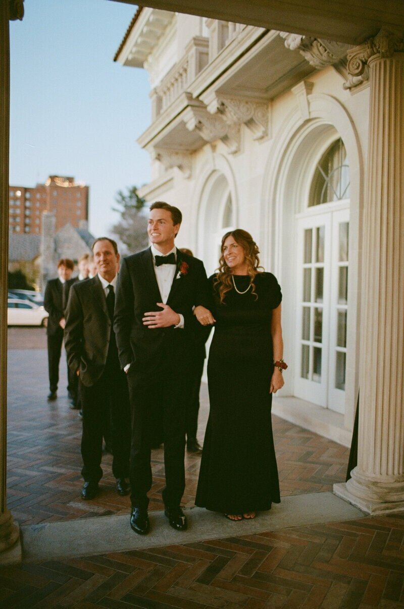 A group of people in formal attire stand in a line on a brick walkway outside a building with ornate columns and arched windows. The foreground shows a man in a tuxedo and a woman in a long black dress walking arm in arm.