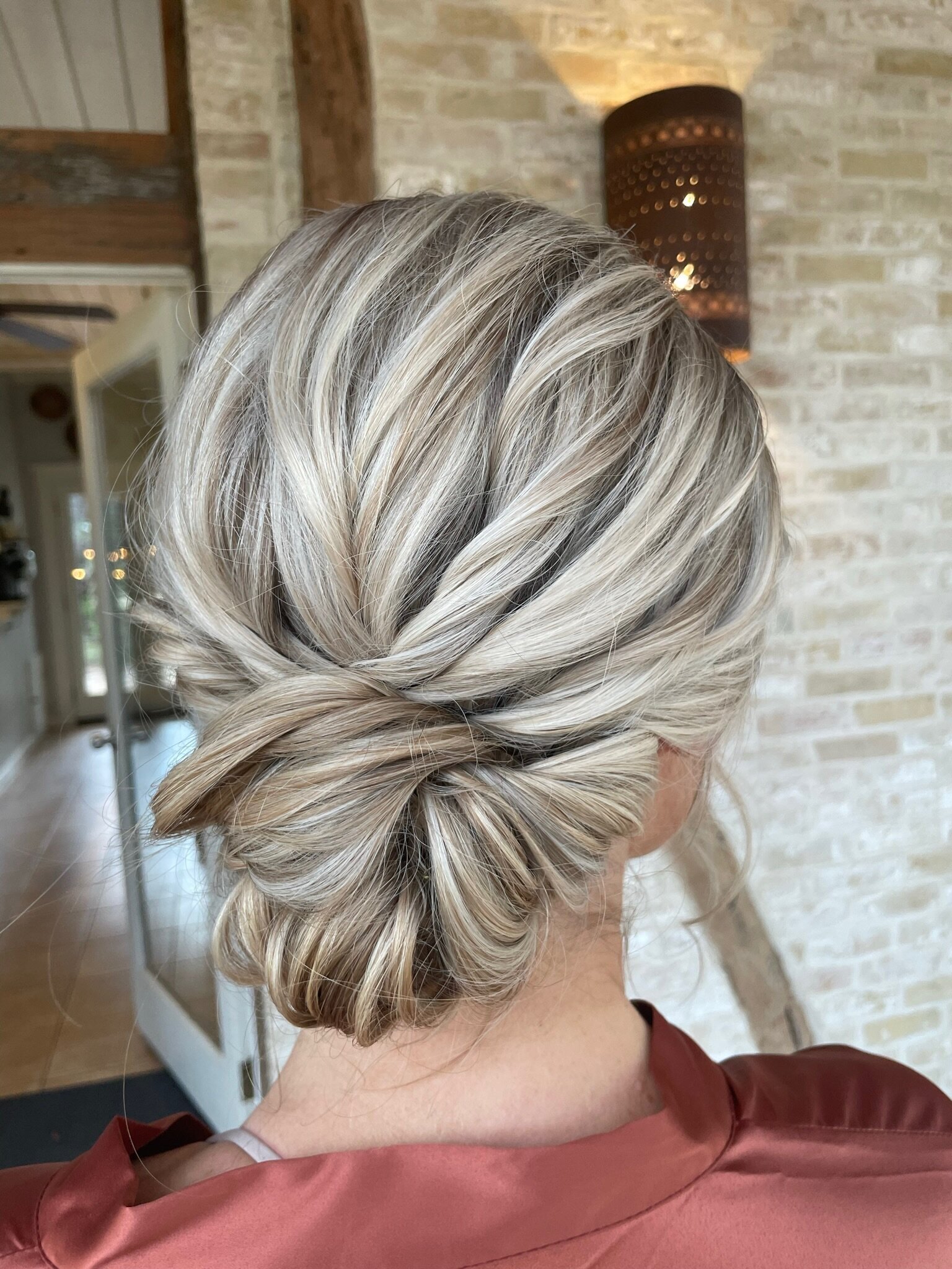 Blonde hair styled in an elegant, twisted low bun against a rustic indoor backdrop showcases what to expect from a wedding makeup artist during a bridal beauty consultation.