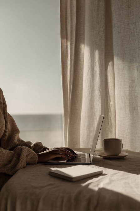 A designer in neutral tones working on a laptop beside a ceramic coffee mug and linen curtains, embodying the slow, intentional creative process at Kathalyst Design Studio.