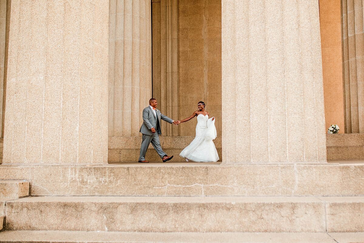The groom, wearing a light gray suit is lead by th ehand by his bride wearing a mermaid lace wedding gown as they walk along the columns at the Parthenon