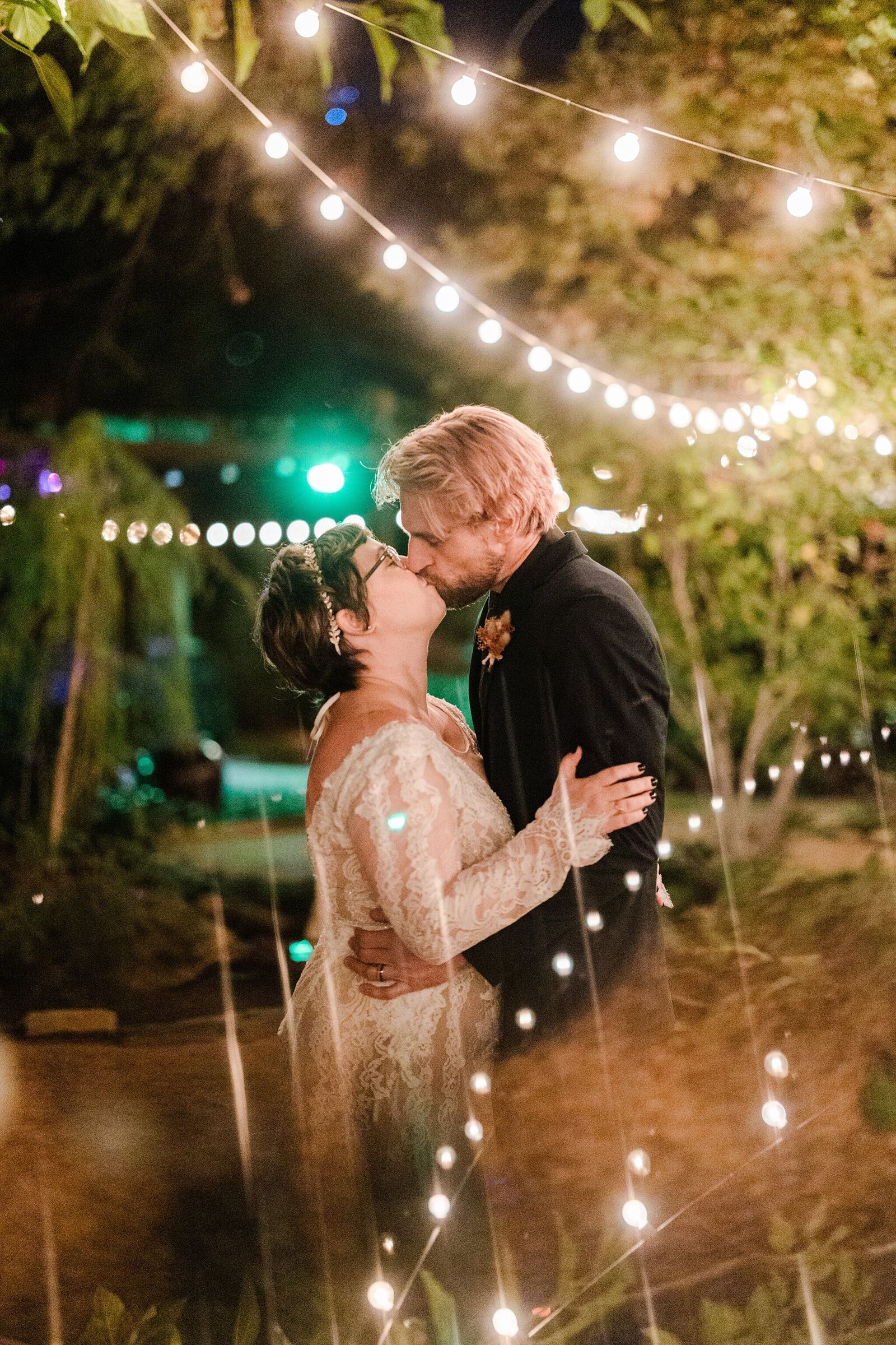 A bride and groom sharing a kiss at a plant nursery venue surrounded by cafe lights and many different plants and trees. The bride is on the left and is wearing a long sleeve lace dress and a flower crown. The groom is on the right and is wearing a black suit and boutonniere.