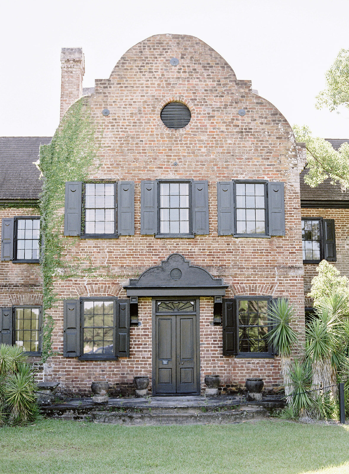 The front of Middleton Place. All brick house with black shutters, black door, and black trim. Palm trees surrounding each side. Photographed by wedding photographers in Charleston Amy Mulder Photography