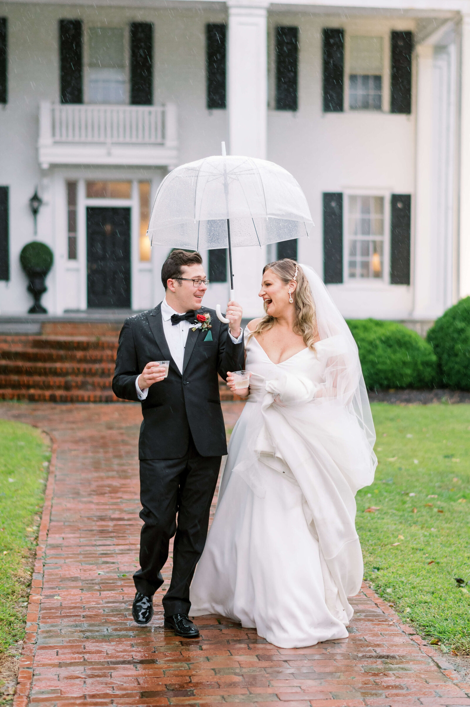 A newly married couple cheers champagne while walking together in the rain outside of a large white Virginia house