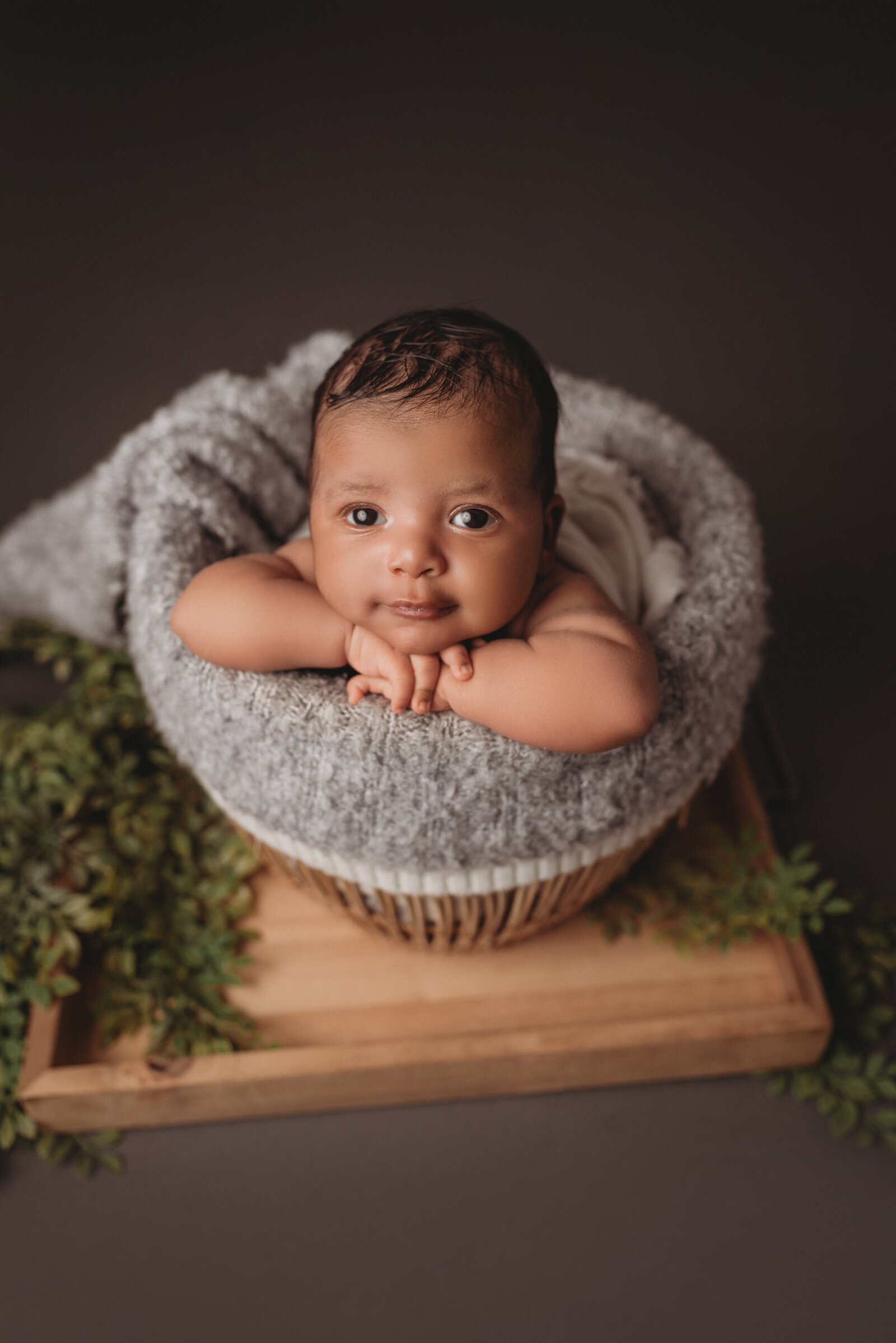 Bright eyed newborn baby boy in rattan basket with fuzzy layers resting chin on hands and looking at the camera, shot on a dark grey backdrop