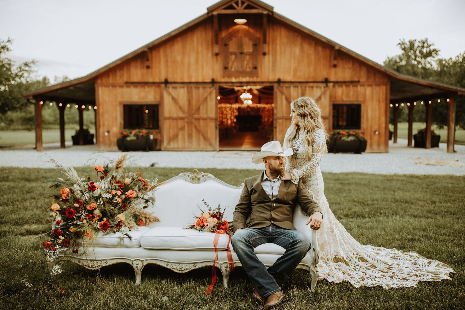 Bride standing beside groom seated on a vintage couch with a floral arrangement, barn in the background