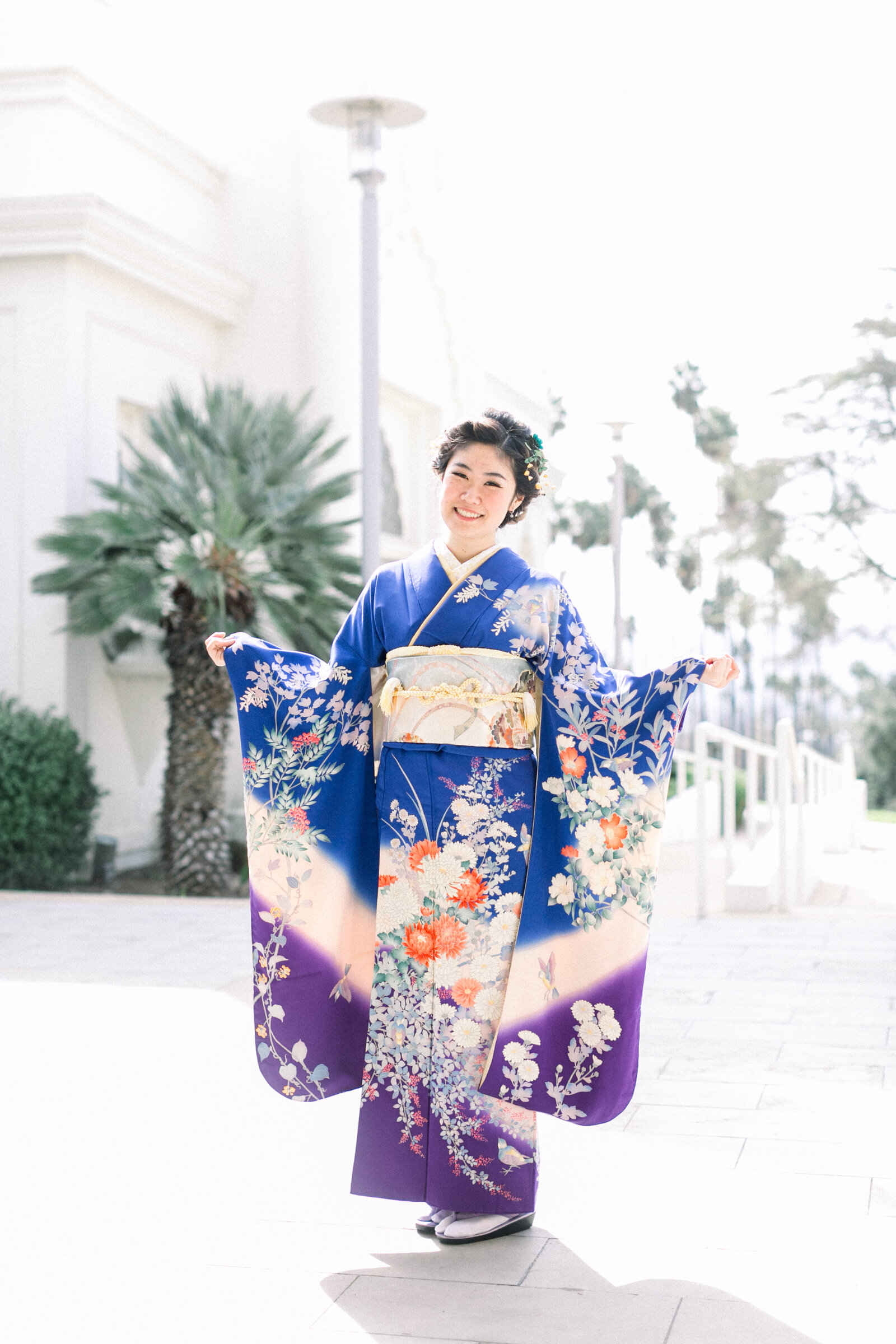Young woman in vibrant blue furisode kimono standing with arms gracefully extended, celebrating Seijinshiki (coming-of-age) at Brand Park Glendale. Kimono rental in Los Angeles | 成人式の振袖を着た女性がブランドパーク（グレンデール）で華やかにポーズ、ロサンゼルスで着物レンタル