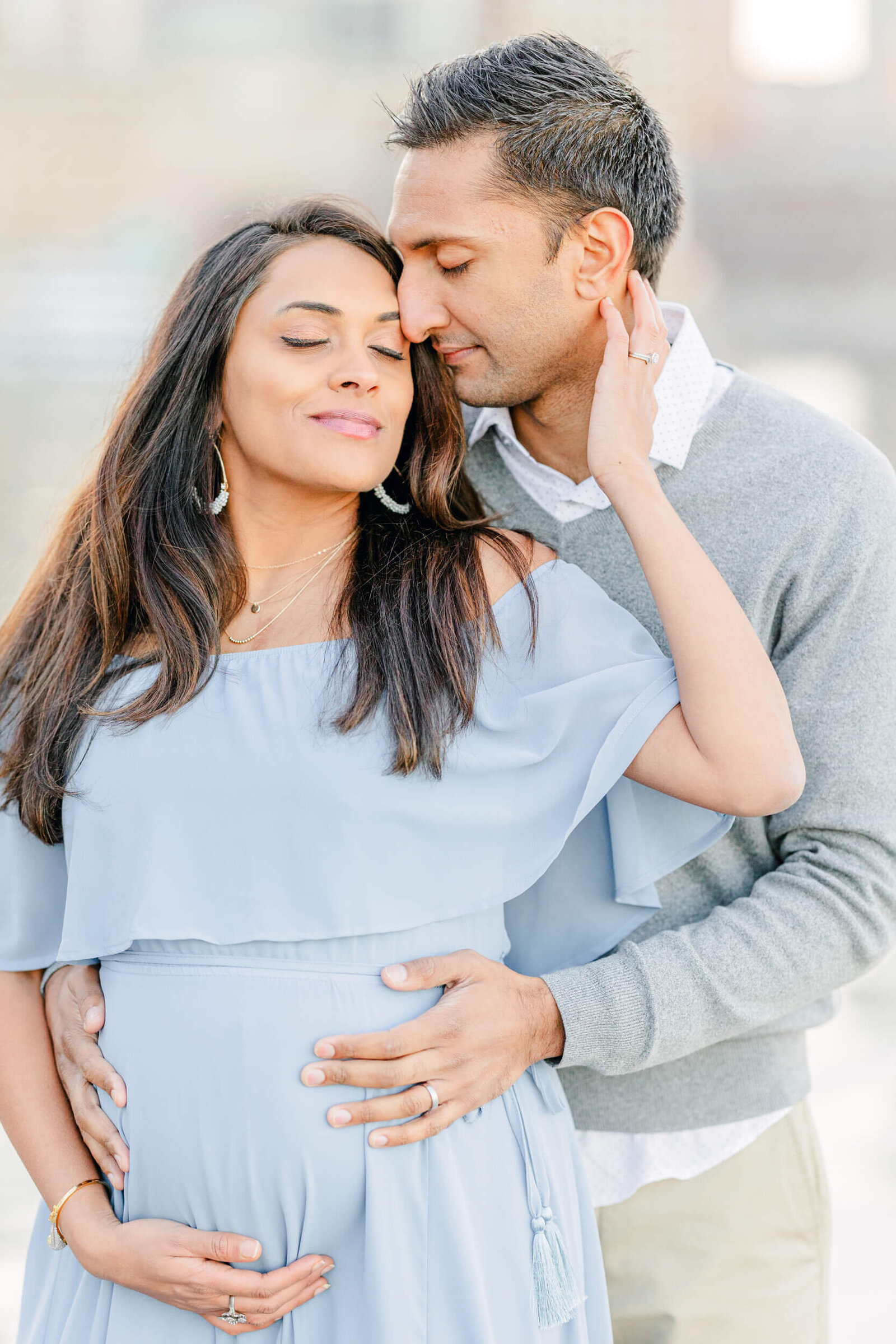 Pregnant woman and her husband stand together with their eyes closed and their hands on her baby bump