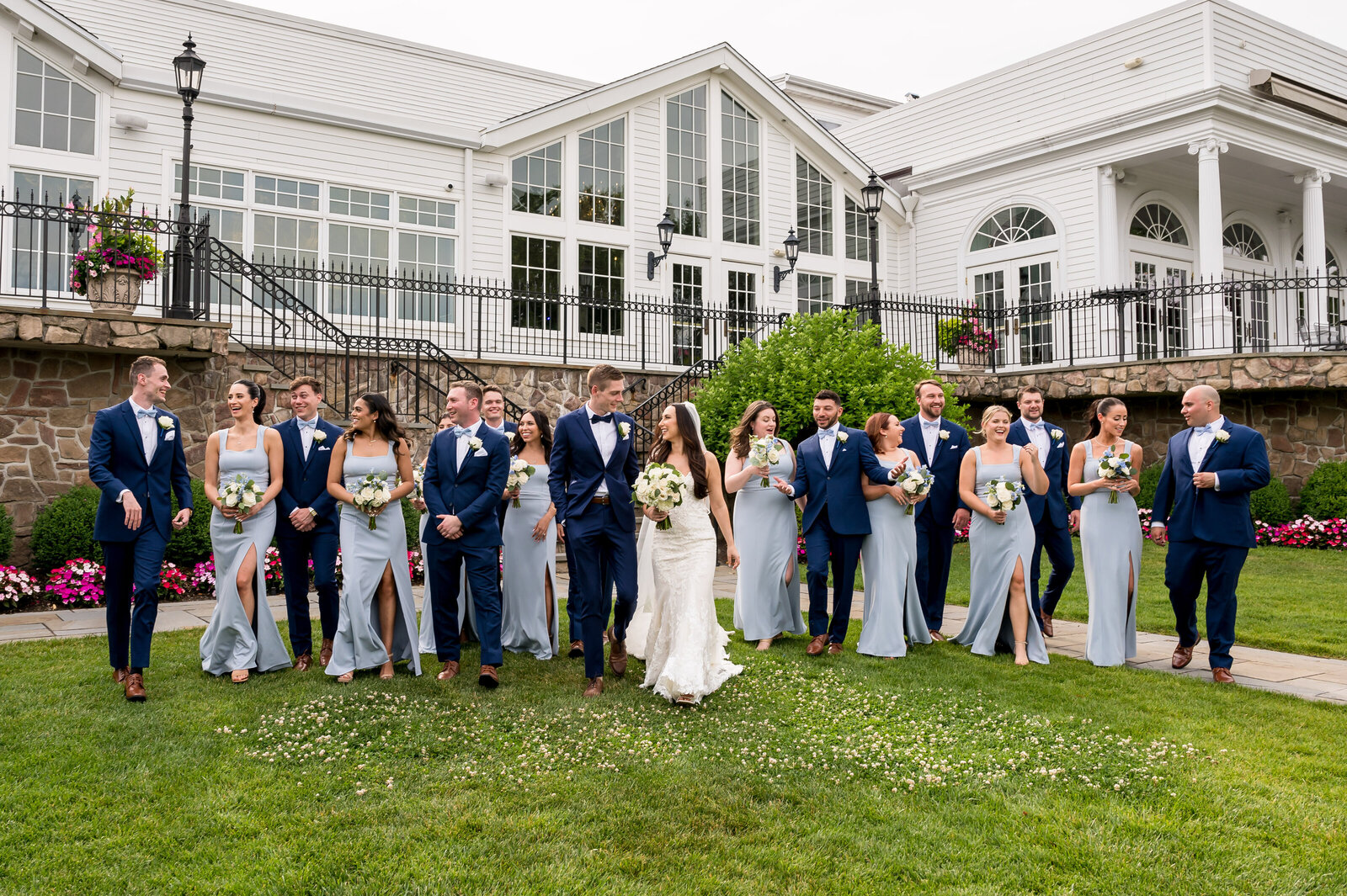 A wedding party in formal attire walks on grass in front of a large building. The group includes bridesmaids in light gray dresses and groomsmen in blue suits.