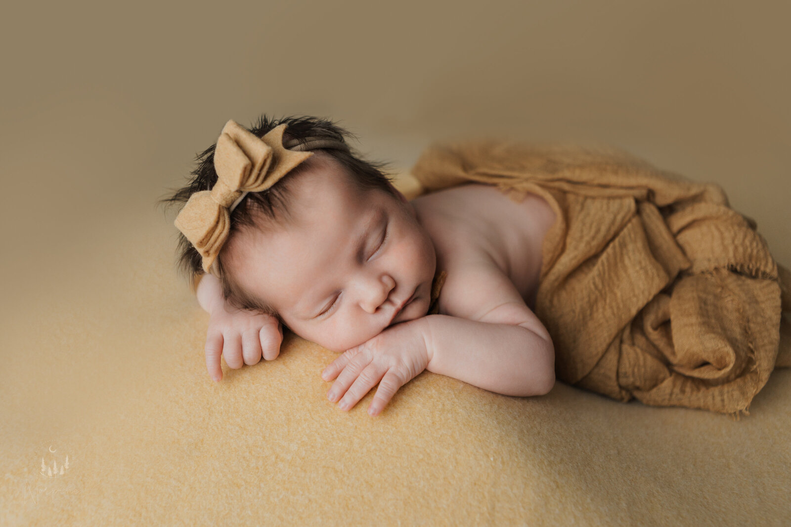 a newborn girl with yellow backdrop, wrap and headband with a bow, laying on her stomach with both arms up near her face, sleeping.