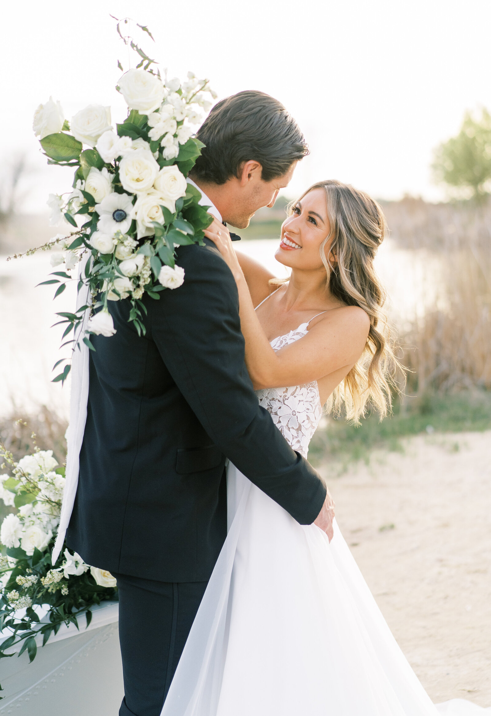 Portrait of a bride in a white wedding gown and bouquet hugging the groom in a black tuxedo in front of a lake