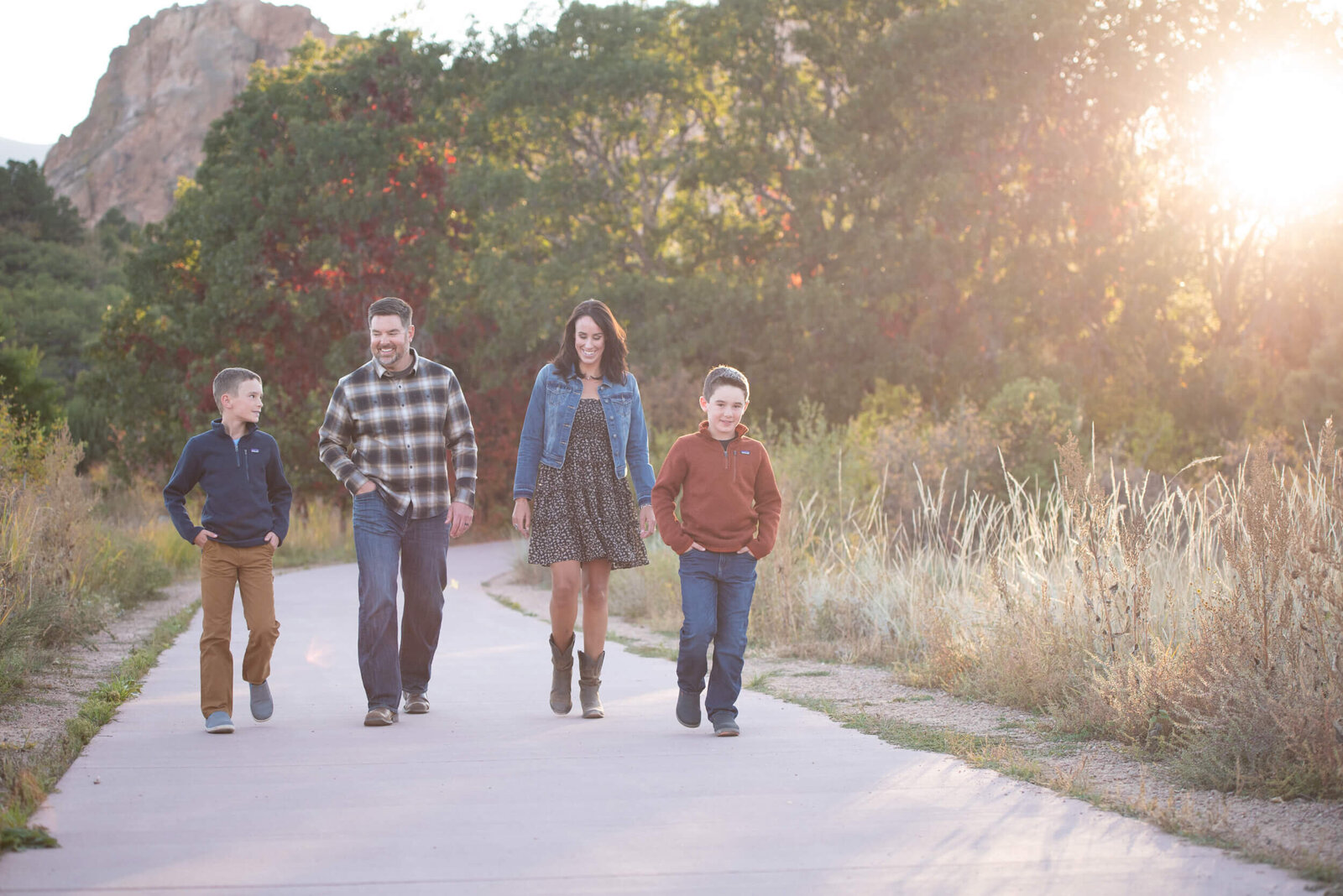 Happy parents walk down a park path with their 2 young sons at sunset with hands in pockets