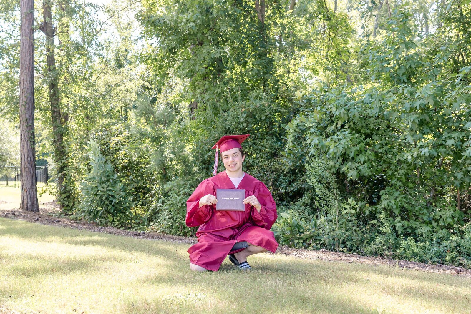 A high school senior in a maroon cap and gown kneeling on the grass and holding a diploma in a park setting in Wilmington, North Carolina. This celebratory photo captures academic achievement, ideal for those seeking outdoor senior photography.