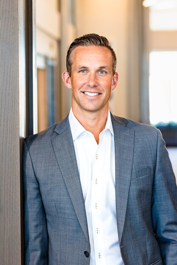 Man in white shirt and gray jacket leans against wall for his headshot.