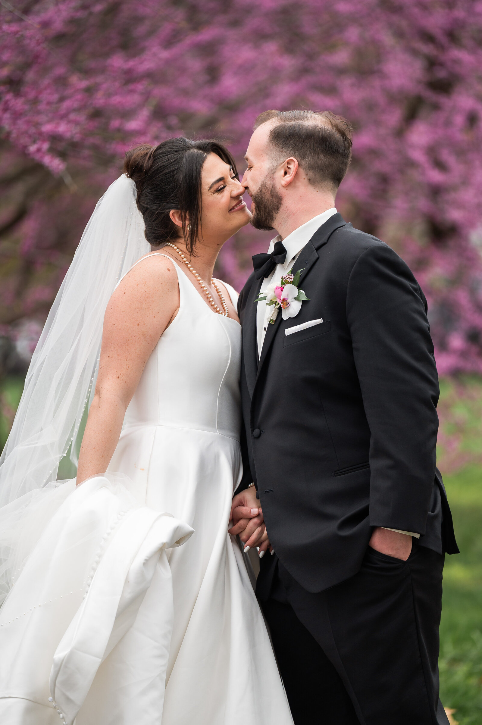A bride and groom stand close, smiling and touching noses, in front of blooming pink trees. The bride wears a white dress and veil; the groom wears a black tuxedo.