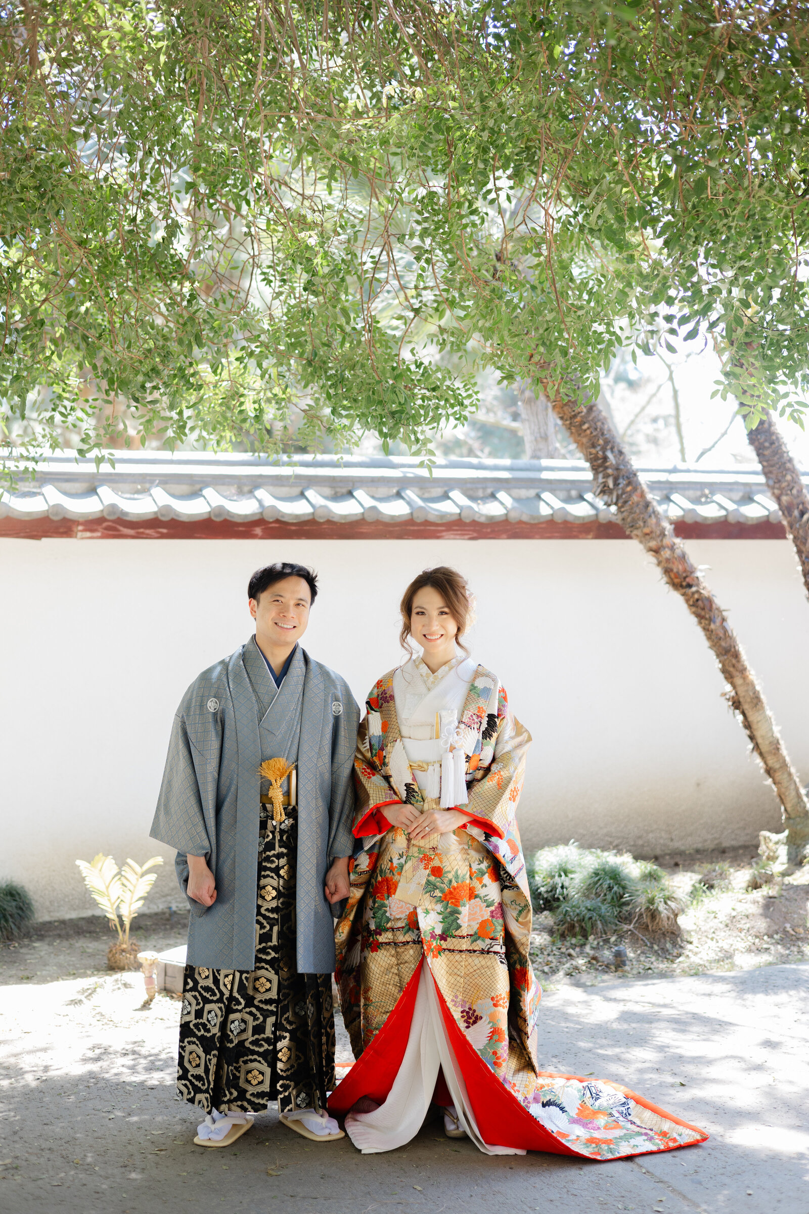 Bride in gold floral uchikake and groom in montsuki standing under trees at Shoseian “Whispering Pine” Tea House, Los Angeles California. Perfect for wedding kimono rental | 書斎庵「ささやき松」の茶室庭園で金色花柄の色打掛花嫁と紋付袴の新郎。ロサンゼルス カリフォルニアの和装着物レンタル