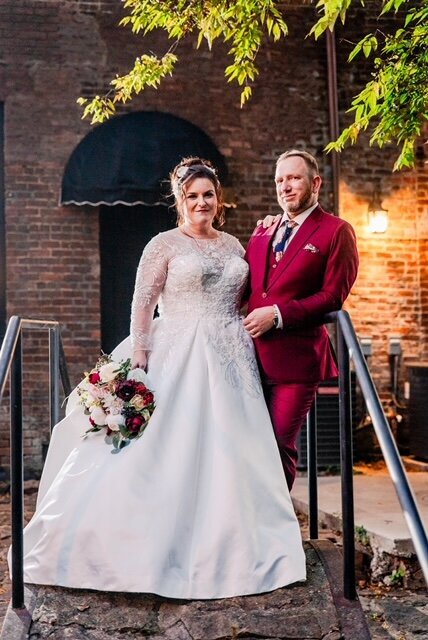 Groom wearing a burgundy tuxedo standing on bridge with bride in her ballgown wedding dress holding a red and blush rose wedding bouquet