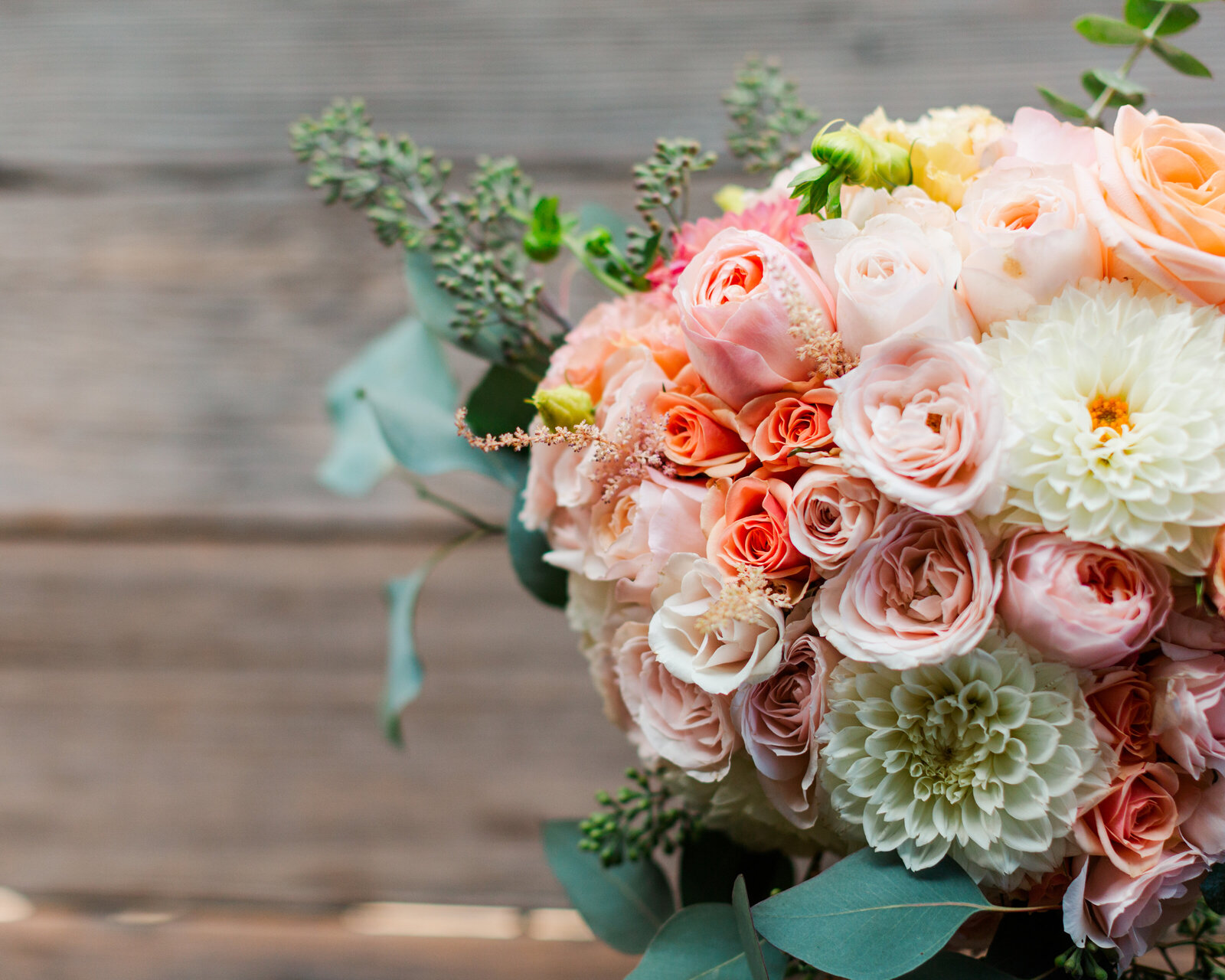 Close up wedding detail photo of a bridal bouquet with a variety of cream, peach, pink, and yellow tone roses, mums, and greenery.