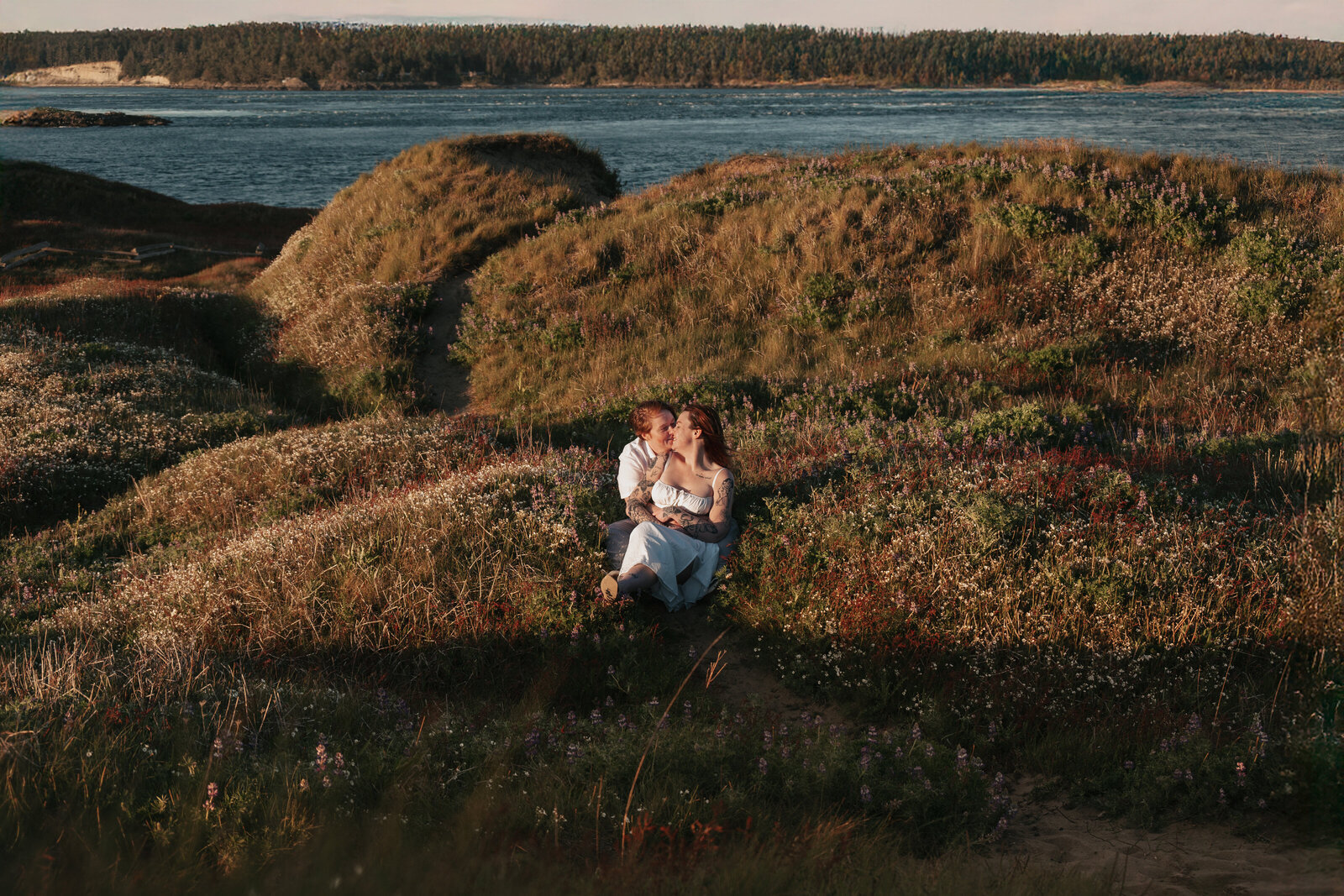 Couple sits on a grassy coastal cliff at golden hour, overlooking the Pacific Ocean at sunset during an intimate engagement session in the San Juan Islands. 