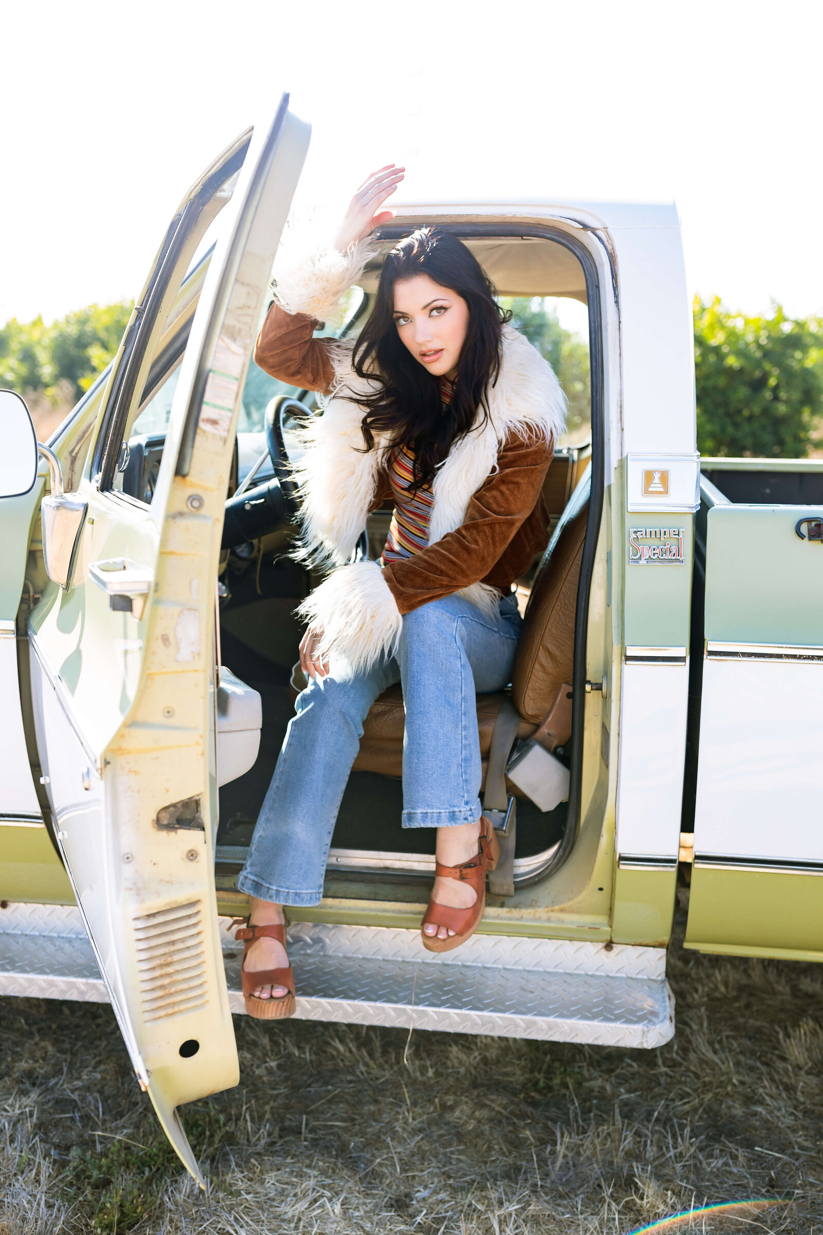 black haired girl wearing brown fur coat sitting on brown leather seats and wearing brown wedged heels