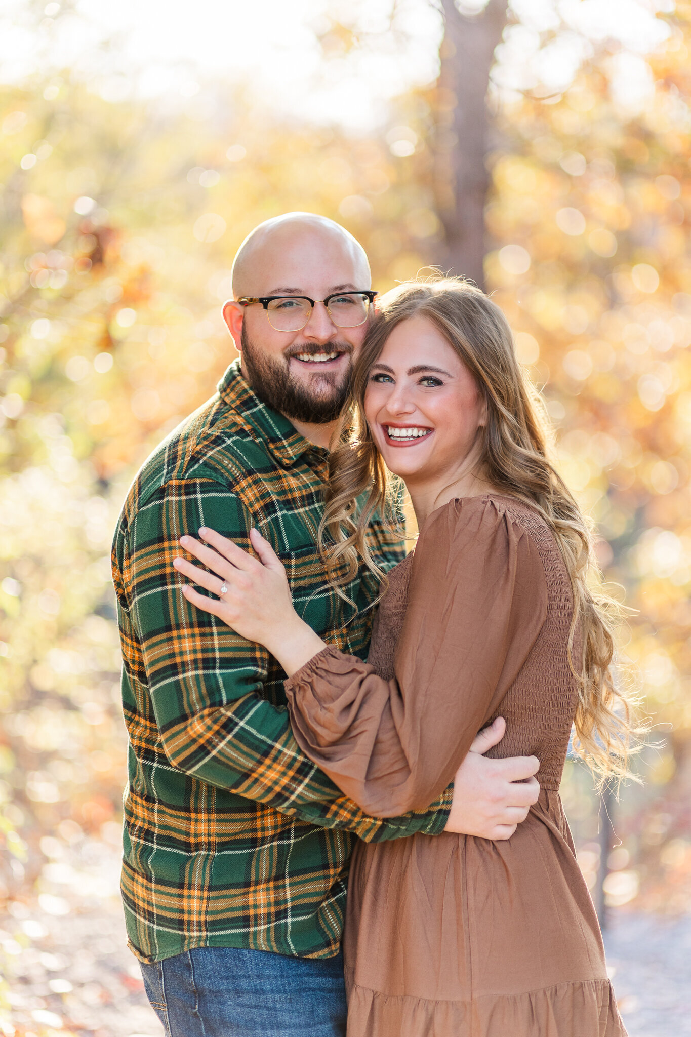 A couple hugs and smiles at camera with fall foliage in background