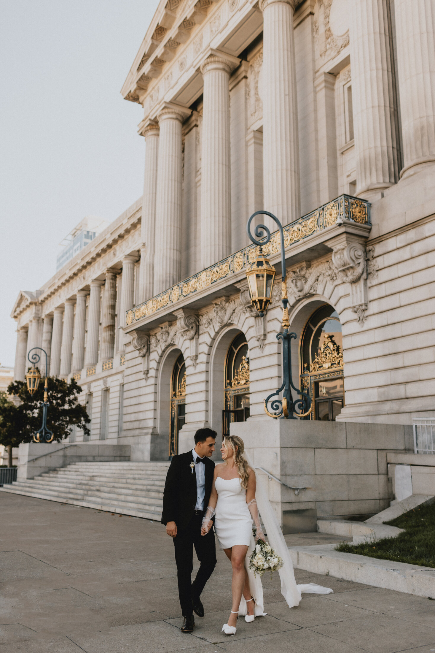 A newly married couple walks hand in hand outside an ornate City Hall building with classical columns and decorative street lamps in San Francisco, the bride in a white dress holding a bouquet and the groom in a black suit