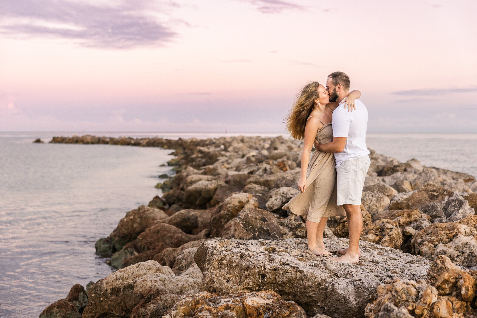A couple kisses while standing on a rock jetty with a beach sunrise in the background