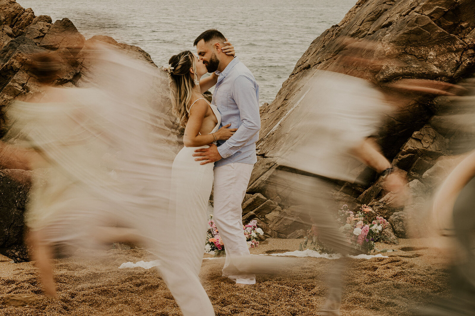 Couple s'embrassant sur la plage, entouré d'amis flous en mouvement, roche et mer en arrière-plan. Photographie prise par Laura en vendée.