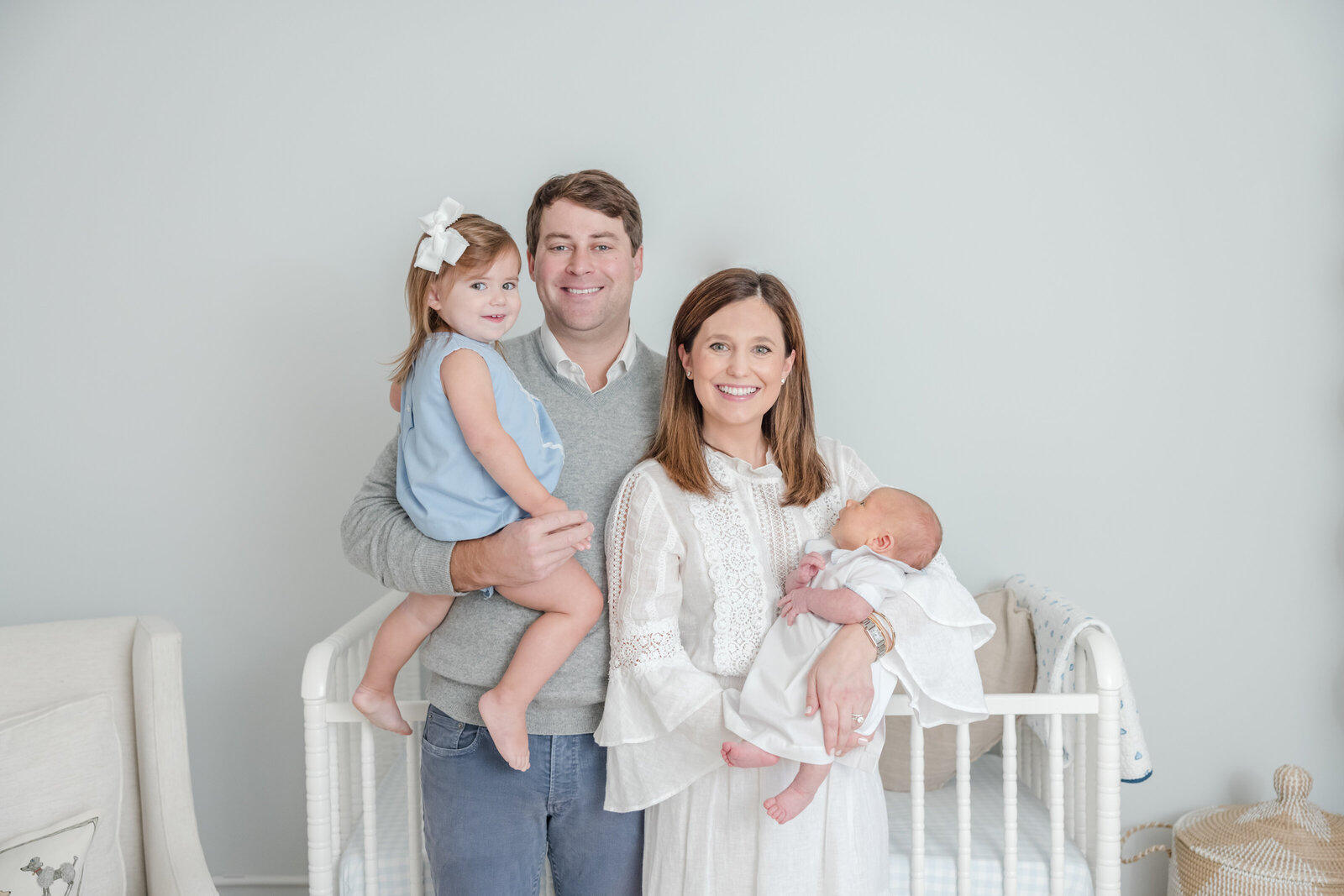 Family of four smiling and standing in front of a crib in the nursery.