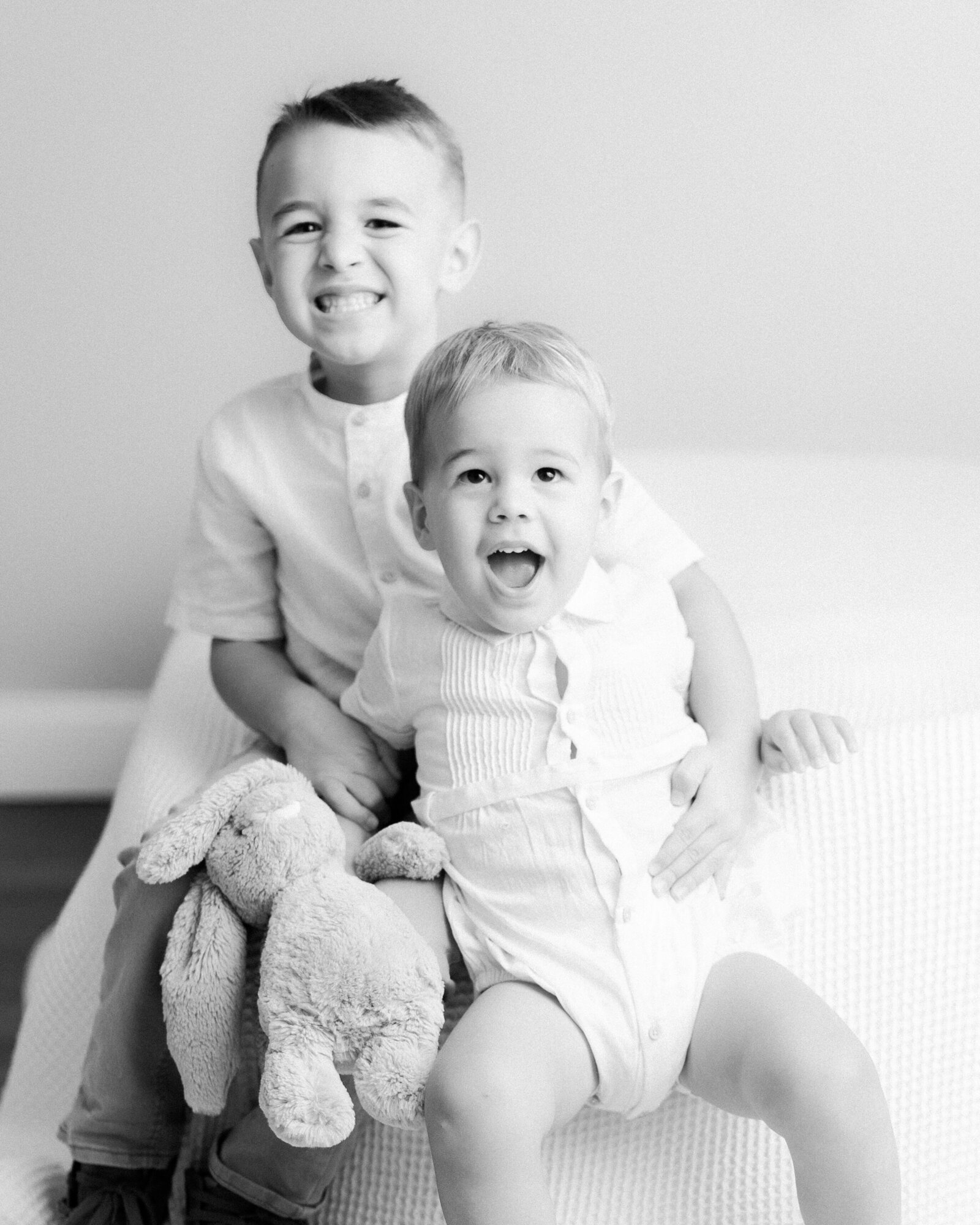 two and four year old brothers sitting on a bed and smiling with their arms around each other by NH newborn photographer Fieldstone Studio.
